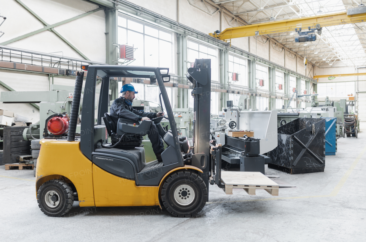 A happy mature man fork lift truck driver lifting pallet in storage warehouse and looking at camera.