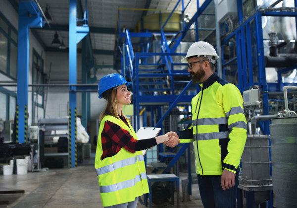 An engineer and industrial worker in uniform shaking hands in large metal factory hall and talking.