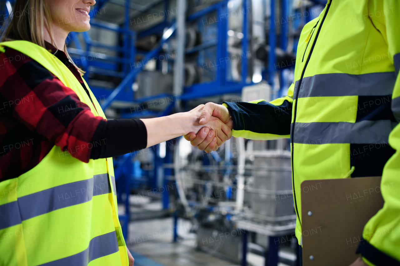 An engineer and industrial worker in uniform shaking hands in large metal factory hall and talking. Close-up.