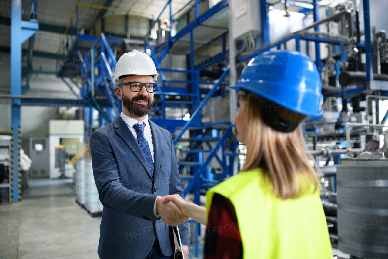 An engineer and industrial worker in uniform shaking hands in large metal factory hall and talking.