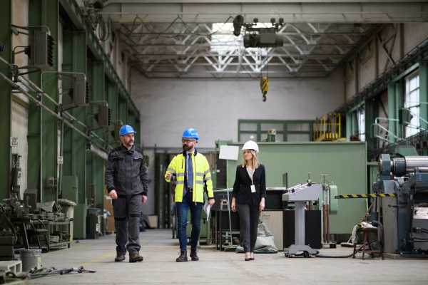 Manager supervisors and industrial worker in a uniform walking in large metal factory hall and talking. Low angle view.