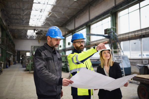 A manager supervisors, engineer and industrial worker in uniform discussing blueprints in large metal factory hall.