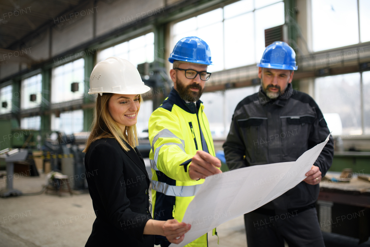 A manager supervisors, engineer and industrial worker in uniform discussing blueprints in large metal factory hall.