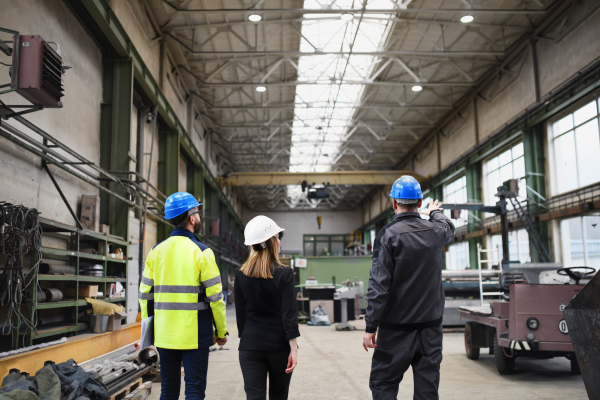 A rear view of manager supervisors and industrial worker in uniform walking in large metal factory hall and talking.