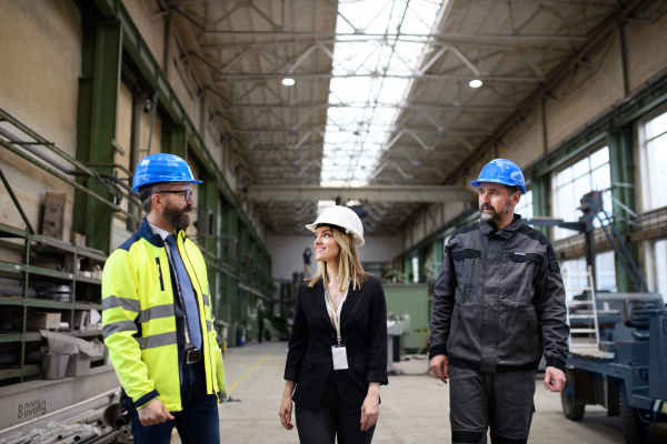 Manager supervisors and industrial worker in a uniform walking in large metal factory hall and talking. Low angle view.