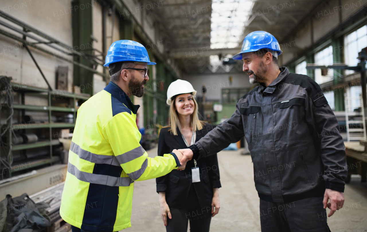 An engineer and industrial worker in uniform shaking hands in large metal factory hall and talking.