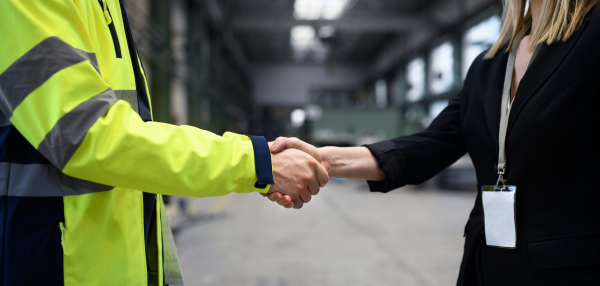 An engineer and industrial worker in uniform shaking hands in large metal factory hall and talking. Close-up.