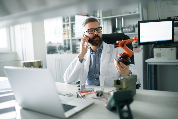 A robotics engineer calling on cellphone and working on desing of modern robotic arm in laboratory office.