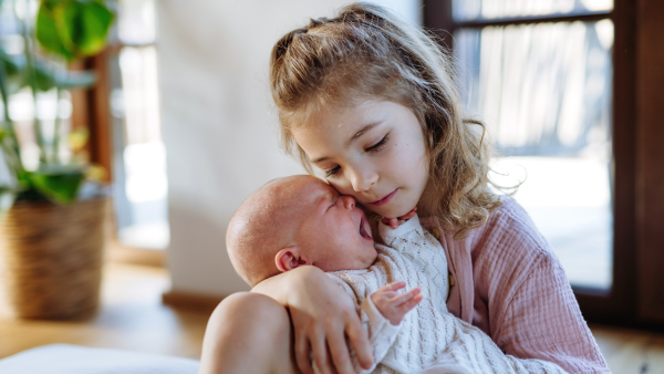 Portrait of big sister soothing newborn sister. Girl taking care of crying little baby. Sisterly love, joy for new family member.