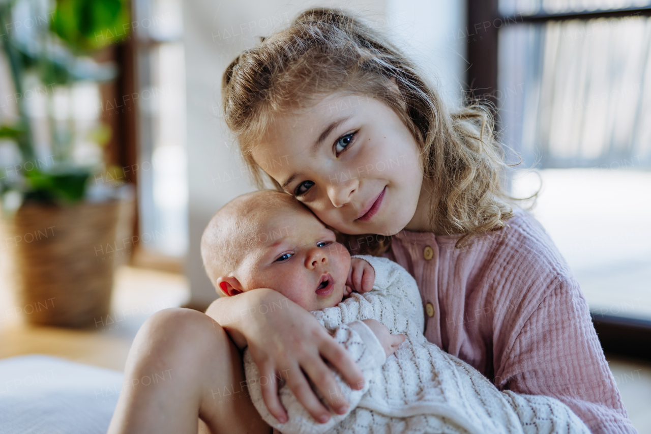 Portrait of big sister holding newborn sister. Girl carefully cuddling little baby. Sisterly love, joy for new family member.