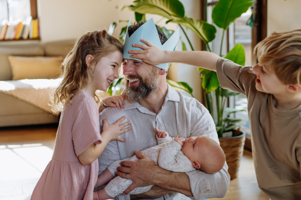 Father with paper crown on head holding newborn baby, enjoying all his kids. New sibling. Unconditional paternal love and Father's Day concept.