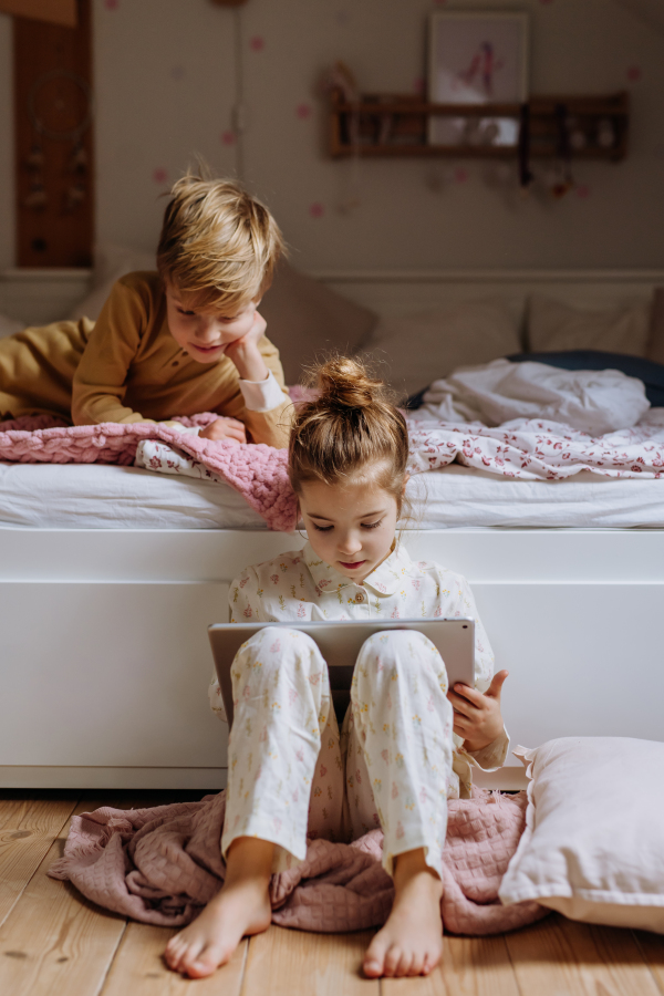 Siblings watching something on tablet, boy lying on bed and cute girl sitting on on floor in bedroom. Children's screen time.