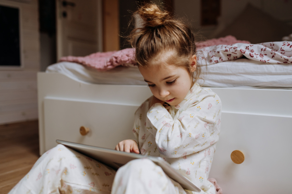 Cute girl sitting on floor in bedroom and watching something on tablet. Children's screen time.