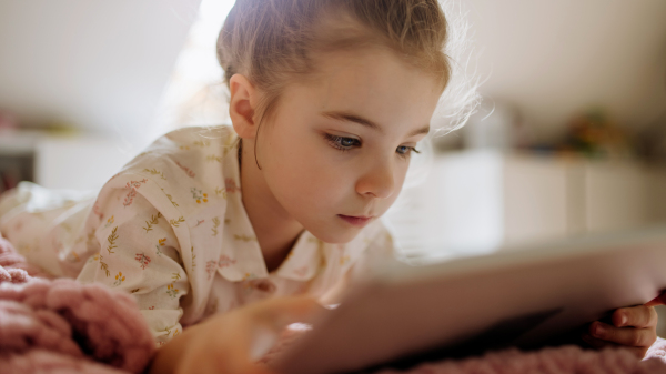 Cute girl lying on bed and watching something on tablet. Children's screen time. Banner with copy space.