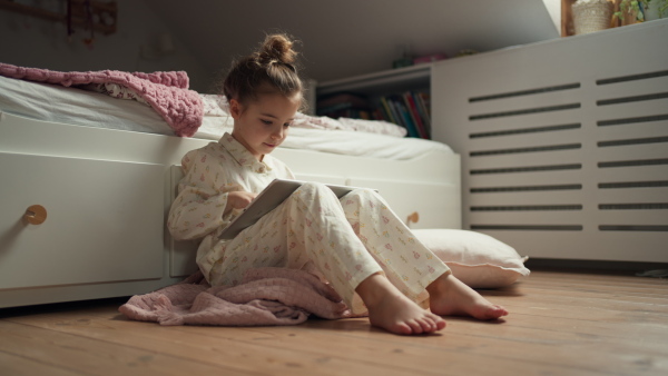 Cute girl sitting on floor in bedroom and watching something on tablet. Children's screen time.