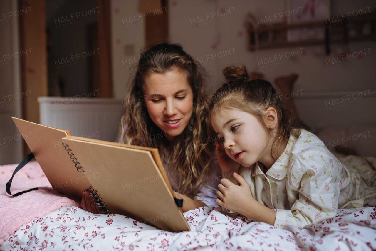 Beautiful mother looking at old family photos in photoalbum with her young daughter, lying on bed in kids room. Calm hygge family moment.