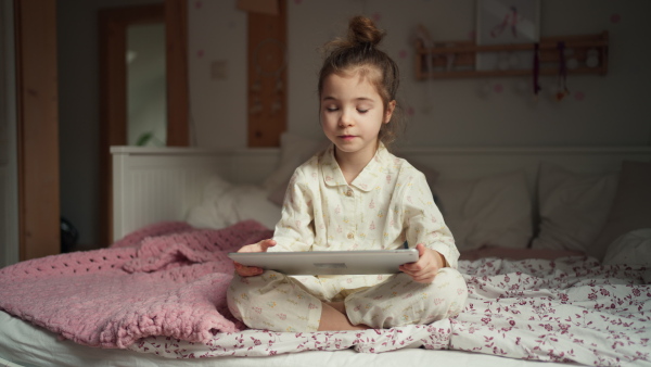 Cute girl sitting on bed in bedroom and watching something on tablet. Children's screen time.