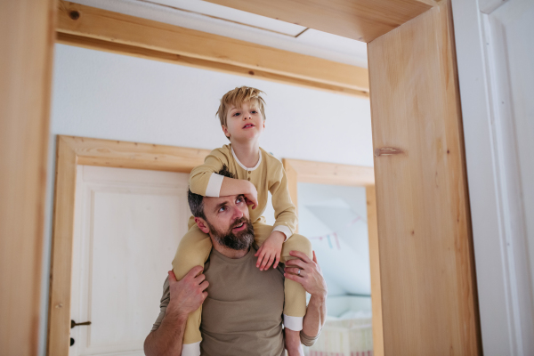 Father carries his son on his shoulders. Boy in pajamas waking up, dad carrying him from bedroom in the morning.