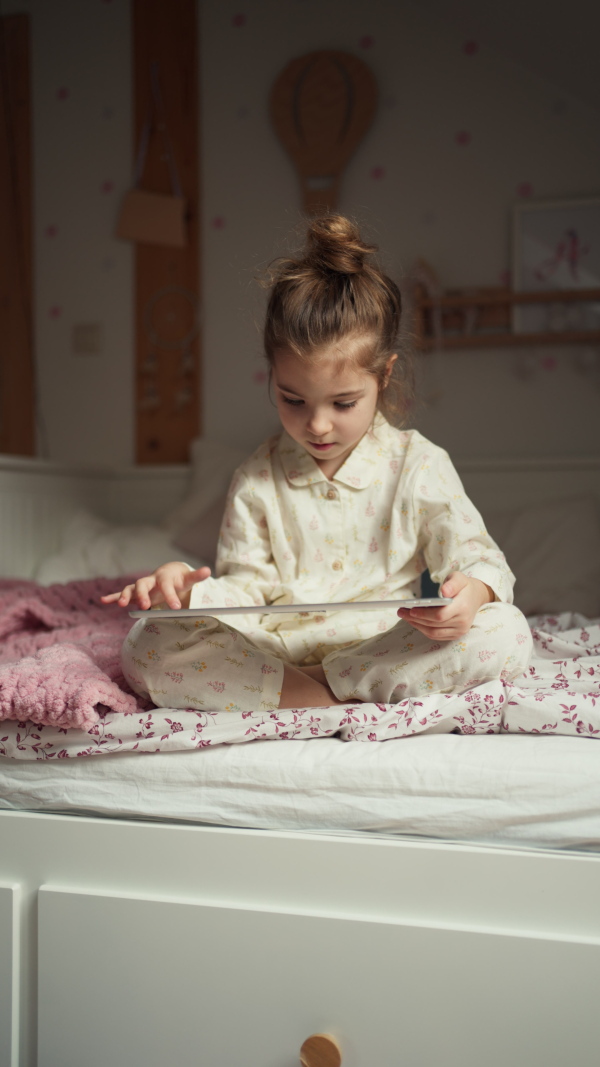 Cute girl sitting on bed in bedroom and watching something on tablet. Children's screen time. Vertical view.