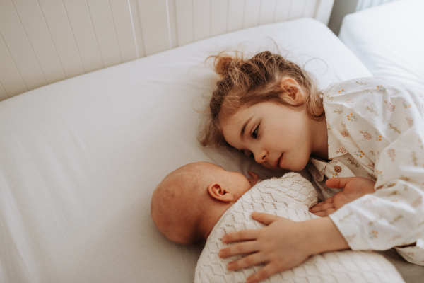 Portrait of big sister cuddling newborn, little baby. Girl lying with her new sibling in bed, closed eyes. Sisterly love, joy for the new family member.