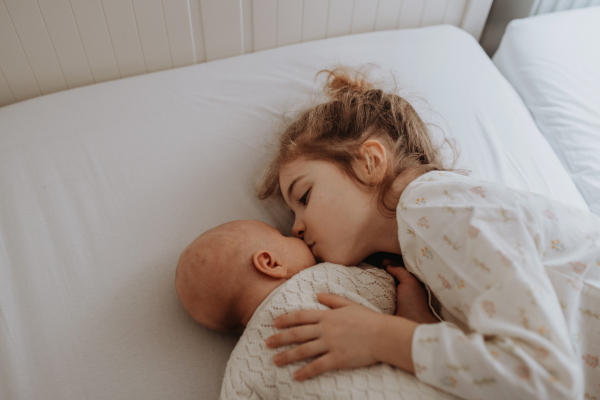 Portrait of big sister cuddling newborn, little baby. Girl lying with her new sibling in bed, kissing baby's cheek. Sisterly love, joy for the new family member.