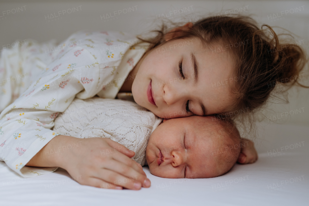 Portrait of big sister cuddling newborn, little baby. Girl lying with her new sibling in bed, closed eyes. Sisterly love, joy for the new family member.