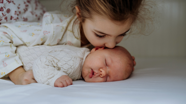 Portrait of big sister cuddling newborn, little baby. Girl kissing her sleeping new sibling in bed. Sisterly love, joy for the new family member.