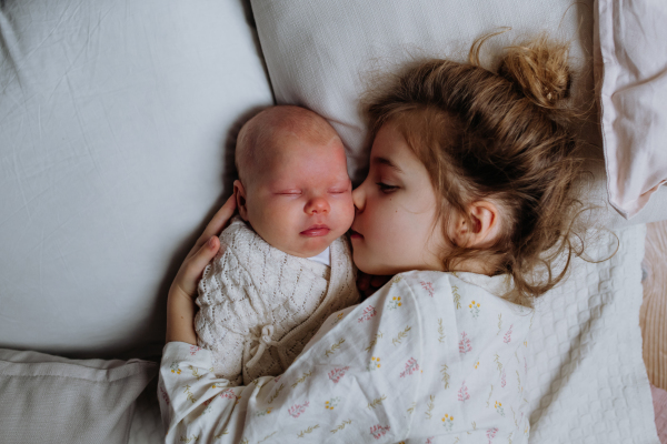 Portrait of big sister cuddling newborn, little baby. Girl lying with her new sibling in bed, closed eyes. Sisterly love, joy for the new family member.