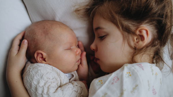 Portrait of big sister cuddling newborn, little baby. Girl lying with her new sibling in bed, closed eyes. Sisterly love, joy for the new family member.