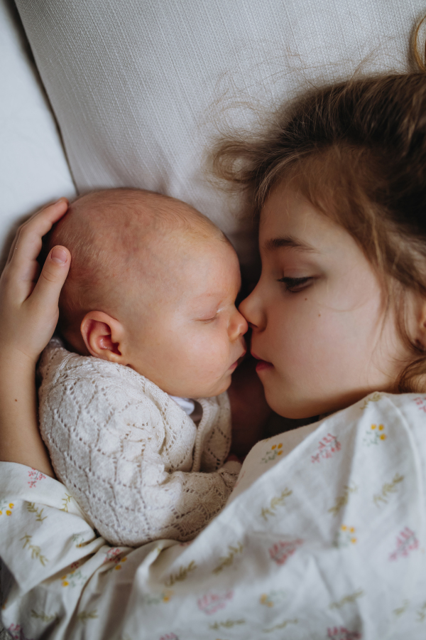 Portrait of big sister cuddling newborn, little baby. Girl lying with her new sibling in bed, closed eyes. Sisterly love, joy for the new family member.