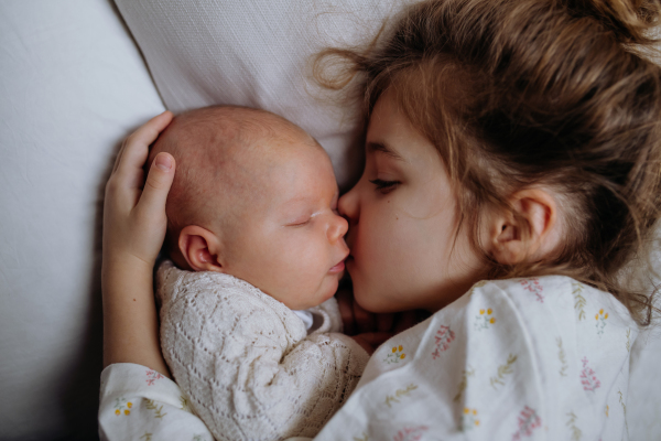 Portrait of big sister cuddling newborn, little baby. Girl lying with her new sibling in bed, kissing baby's cheek. Sisterly love, joy for the new family member.