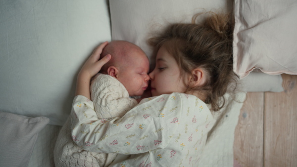 Portrait of big sister cuddling newborn, little baby. Girl lying with her new sibling in bed, closed eyes. Sisterly love, joy for the new family member.