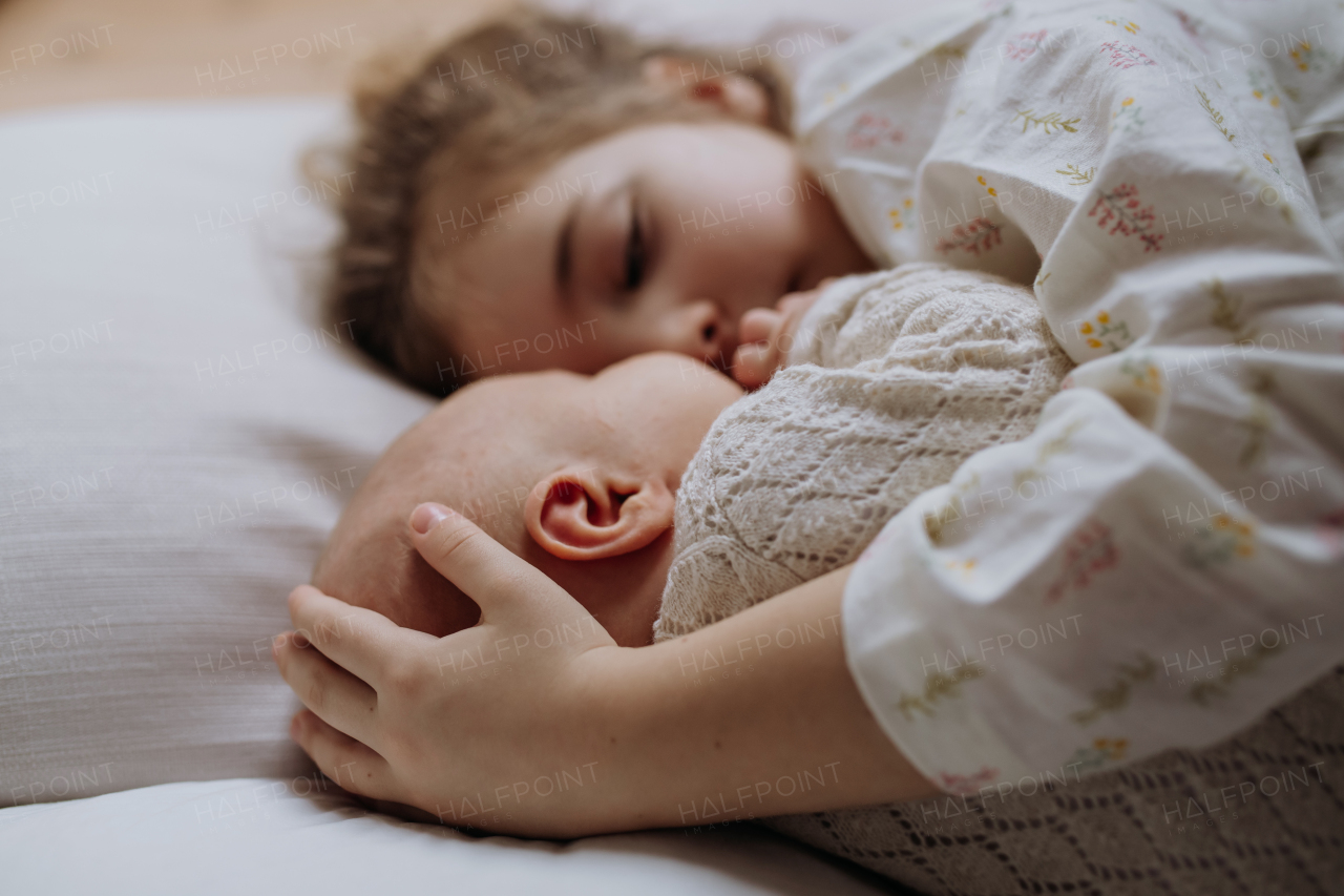 Portrait of big sister cuddling newborn, little baby. Girl lying with her new sibling in bed, closed eyes. Sisterly love, joy for the new family member.