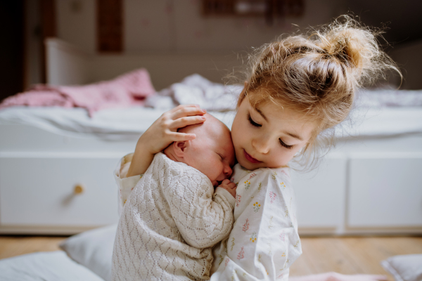 Portrait of big sister holding newborn sister. Girl carefully cuddling little baby. Sisterly love, joy for new family member.