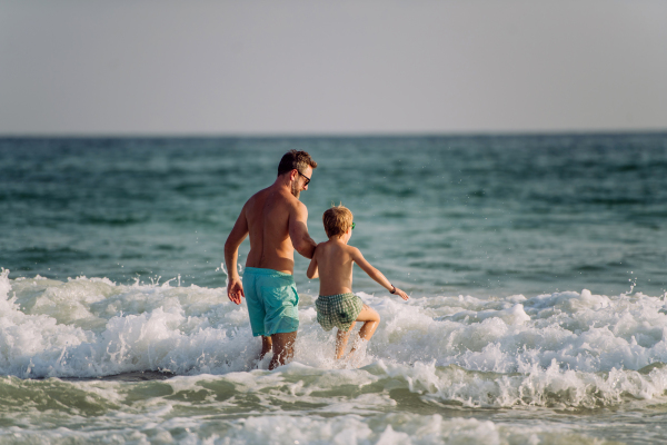 Father with his little son entering in sea, enjoying summer vacation.