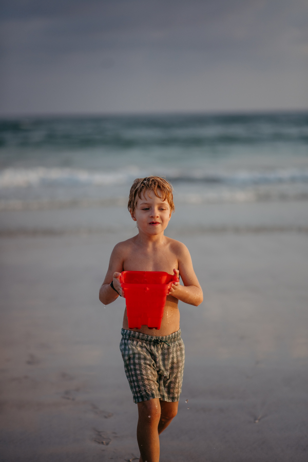 Little boy in swimsuit with bucket full of water running out of sea, enjoying summer holiday.