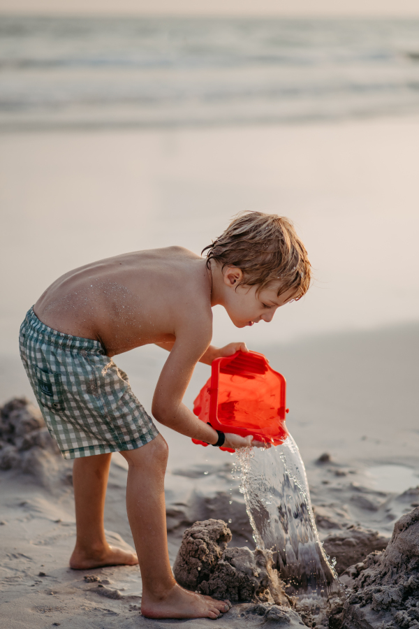 Little boy in swimsuit playing with bucket near the sea, enjoying summer holiday.