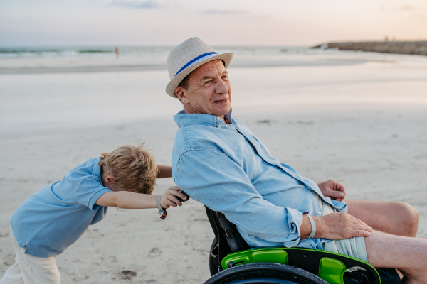 Little boy pushing his granfather on the wheelchair, enjoying sea together.