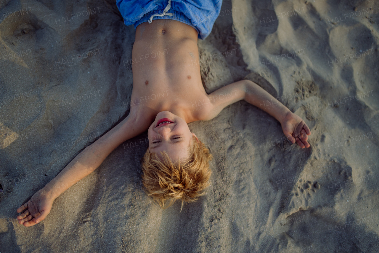 Top view of little playful boy lying in sand on the beach.