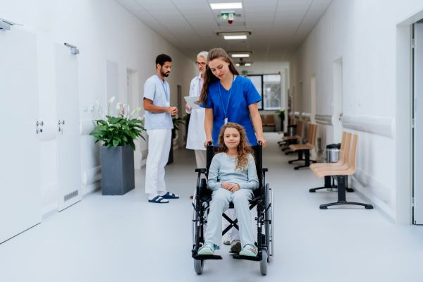 Young nurse pushing little girl on wheelchair at a hospital corridor.