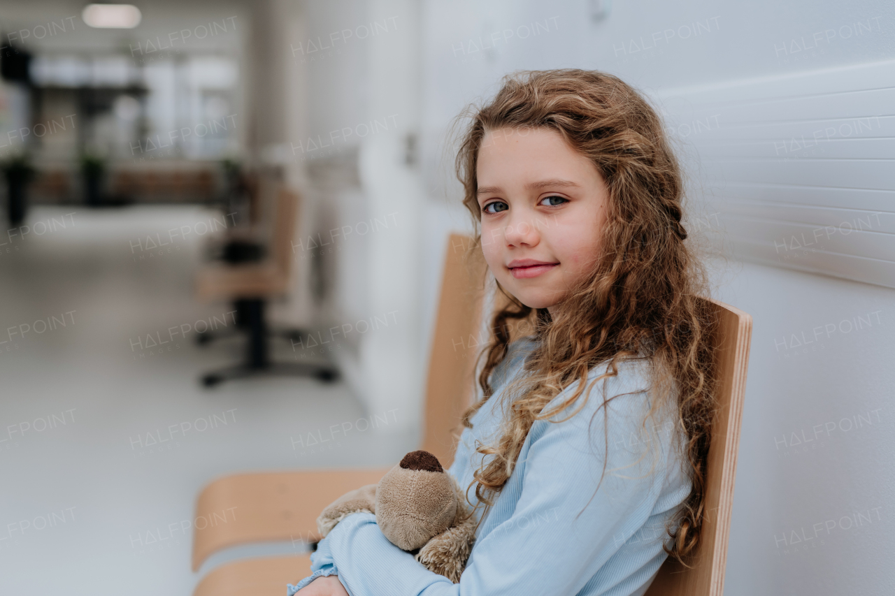 Portrait of little girl with a teddy bear sitting and waiting at hospital corridor.