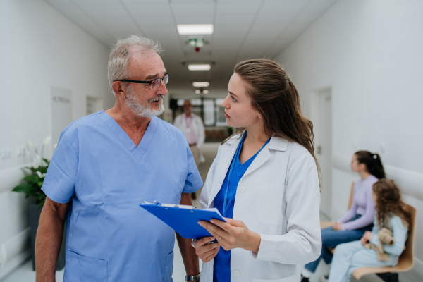 Older doctor giving advise to his younger colleague, discussing at a hospital corridor. Health care concept.