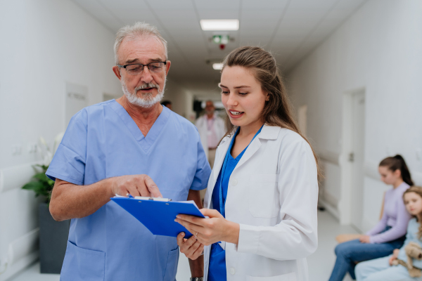 Older doctor giving advise to his younger colleague, discussing at a hospital corridor. Health care concept.