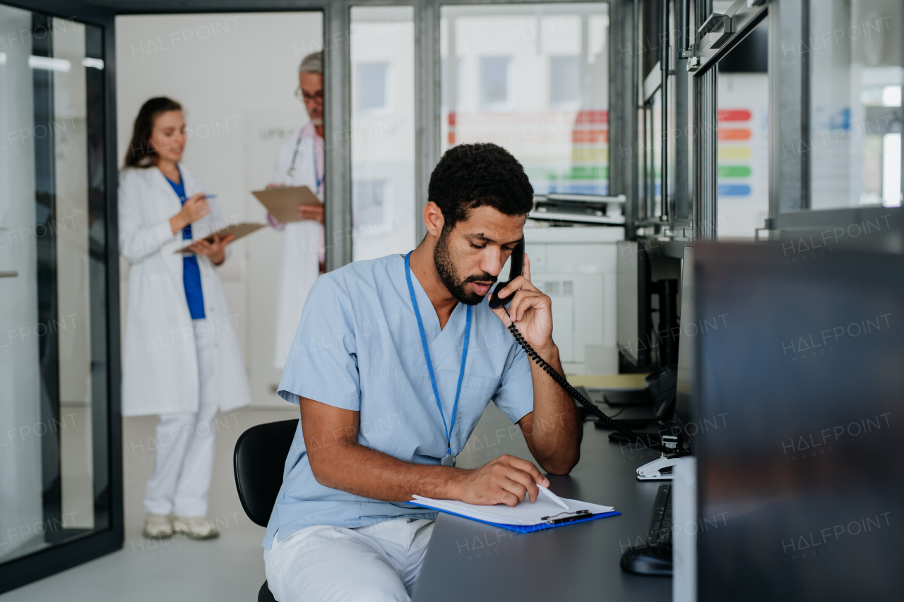 Young multiracial nurse sitting at reception and calling,his colleagues discussing in the background.