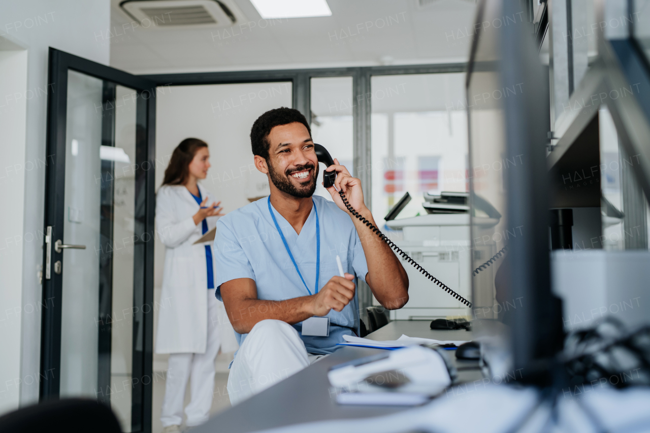 Young multiracial nurse sitting at reception and calling,his colleagues discussing in the background.