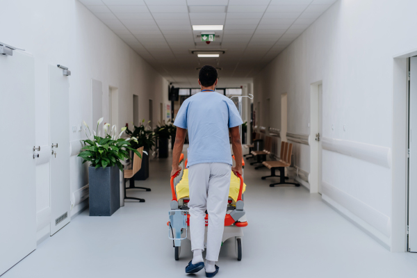 Rear view of caregiver pushing hospital bed at a corridor.