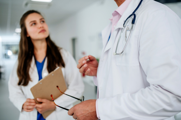 Older doctor giving advise to his younger colleague, discussing at a hospital corridor. Health care concept.