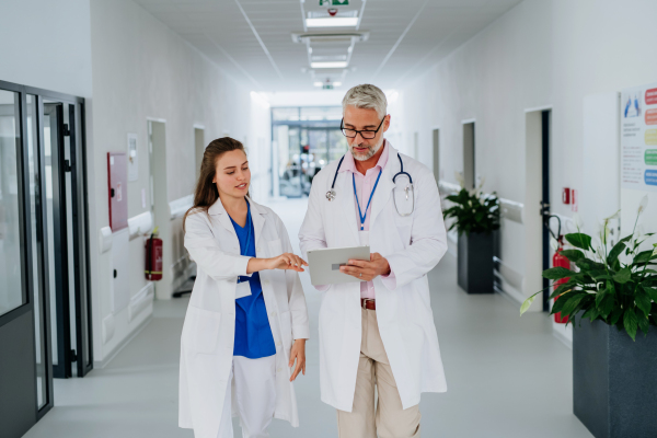 Older doctor giving advise to his younger colleague, discussing at a hospital corridor. Health care concept.