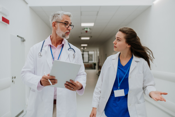 Older doctor giving advise to his younger colleague, discussing at a hospital corridor. Health care concept.