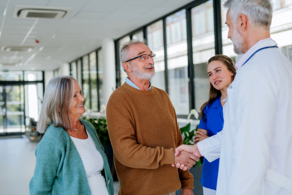 Mature doctor and his young colleaguetalking with his senior patients at hospital corridor.